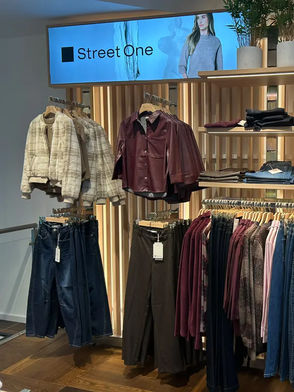 Sous l'enseigne Street One : une veste écrue à carreaux et un blouson de cuir bordeaux, au-dessus d'un portant de jeans et de pantalons