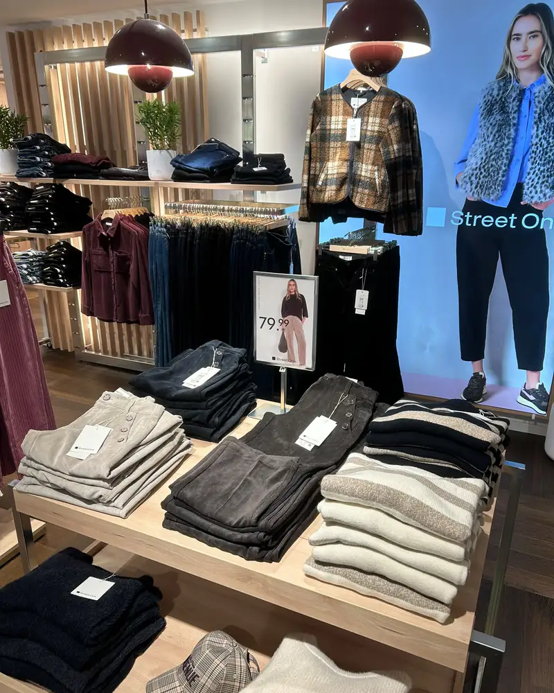 Sur la table, des pantalons en velours beige, gris et noir et des pulls rayés, devant le mur de jeans et le panneau lumineux Street One